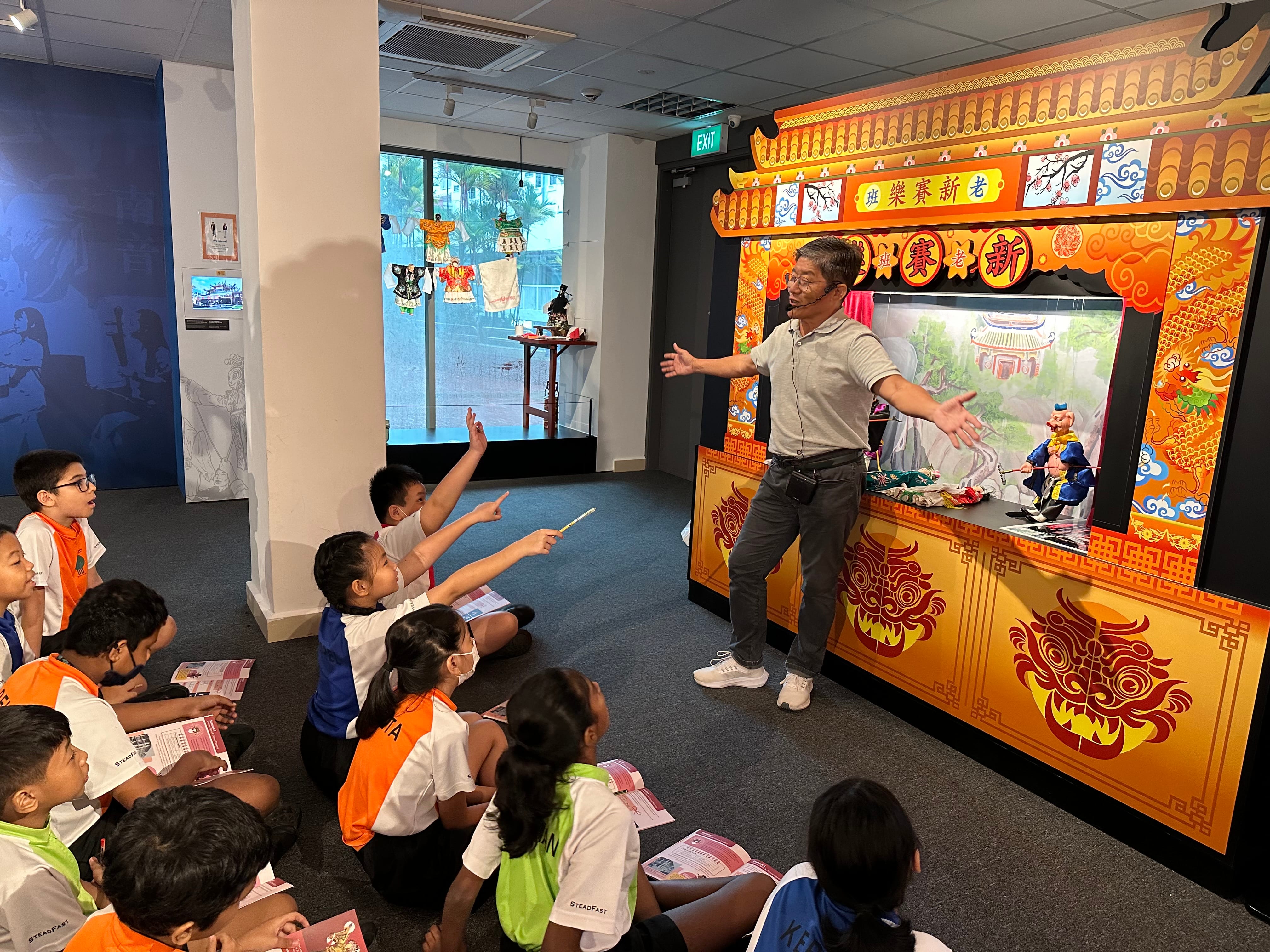 Man speaks with arms wide by a Chinese puppet stage as seated children raise hands and point.